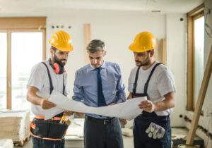Three ADU contractors in Los Angeles wearing hard hats and safety vests examining blueprints at a residential construction site.
