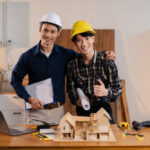 Two ADU contractors in Los Angeles wearing hard hats stand by a table displaying a model of a house, discussing construction plans