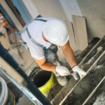 A man in a hard hat and white shirt mixes cement for concrete steps on a front porch.