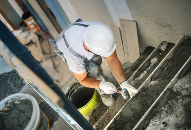A man in a hard hat and white shirt mixes cement for concrete steps on a front porch.