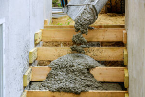 A cement mixer is actively pouring concrete into a wooden stairway, demonstrating the construction of concrete steps for a porch.