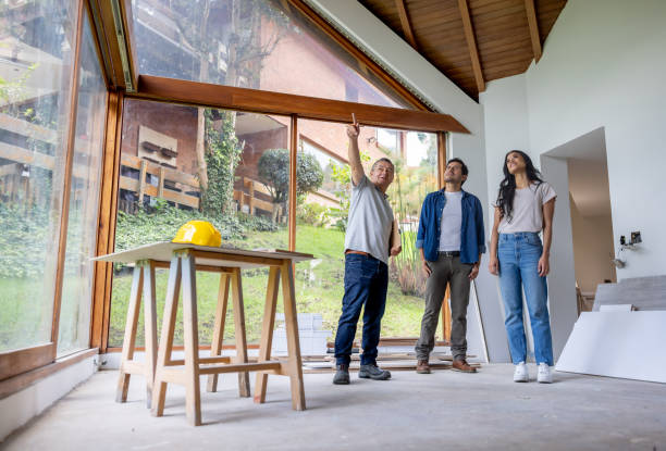 Three individuals stand in an unfinished house, discussing home remodeling ideas amidst exposed walls and construction materials.